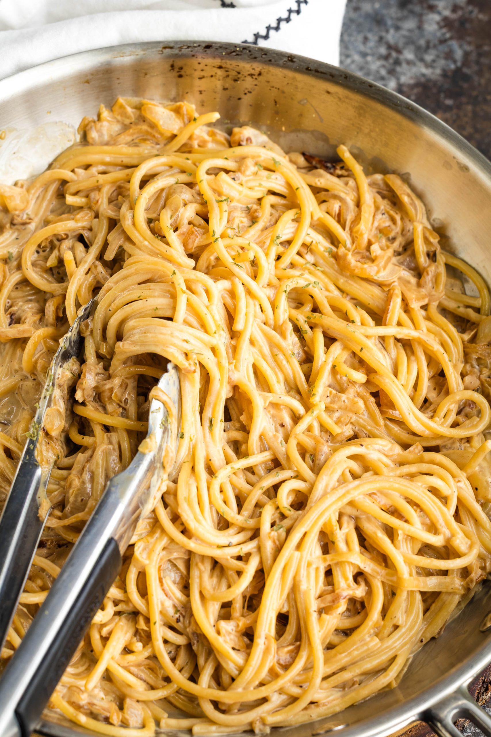 A close-up of creamy caramelized onion pasta being stirred with metal tongs in a stainless steel pan.