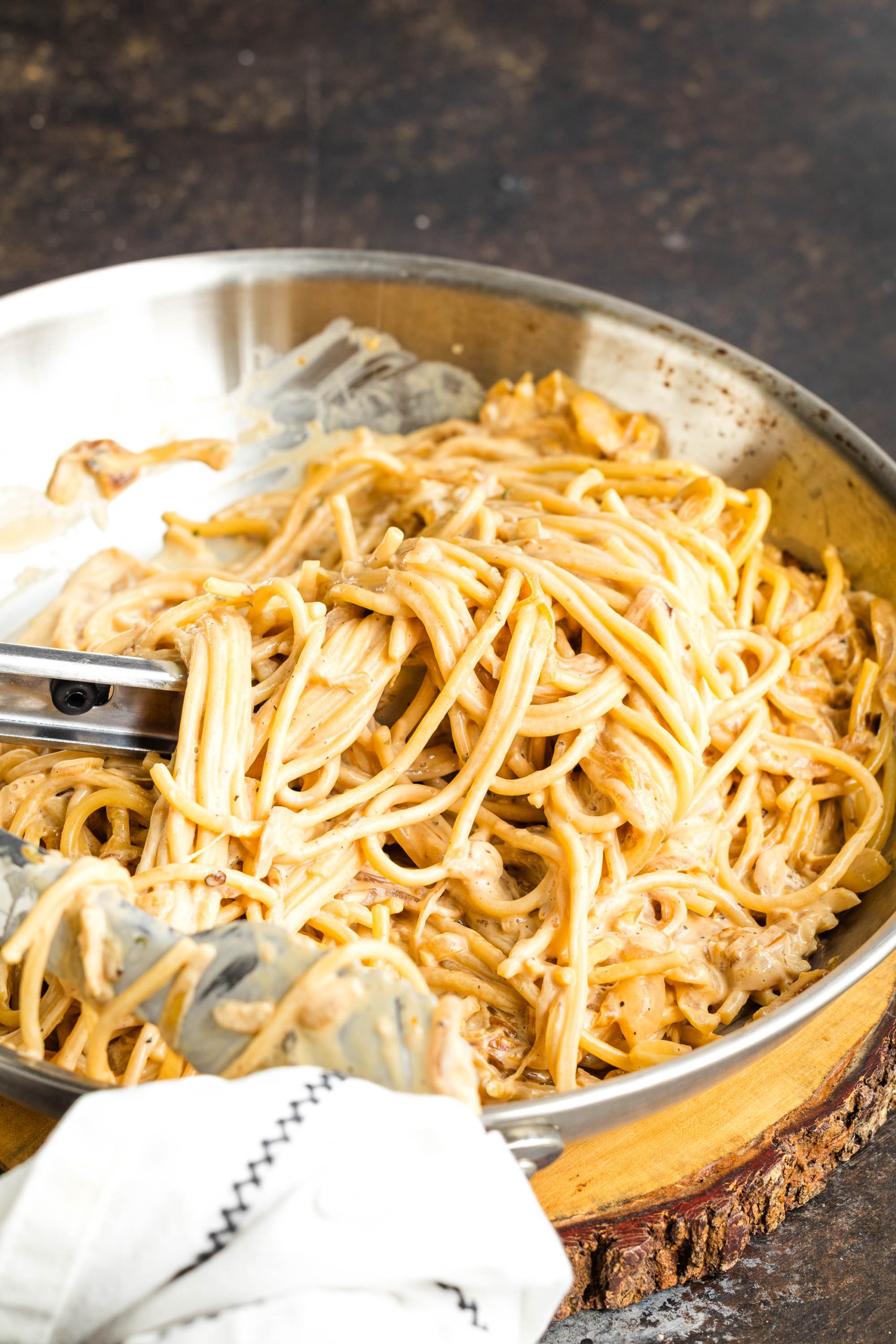 A pan of creamy caramelized onion pasta mixed with a luscious sauce, with metal tongs and a white cloth napkin resting on the side.