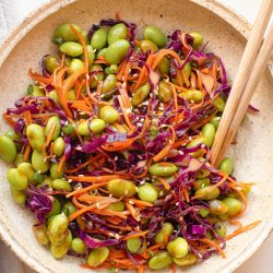 A bowl of salad with edamame, shredded carrots, red cabbage, and sesame seeds, mixed with wooden chopsticks.