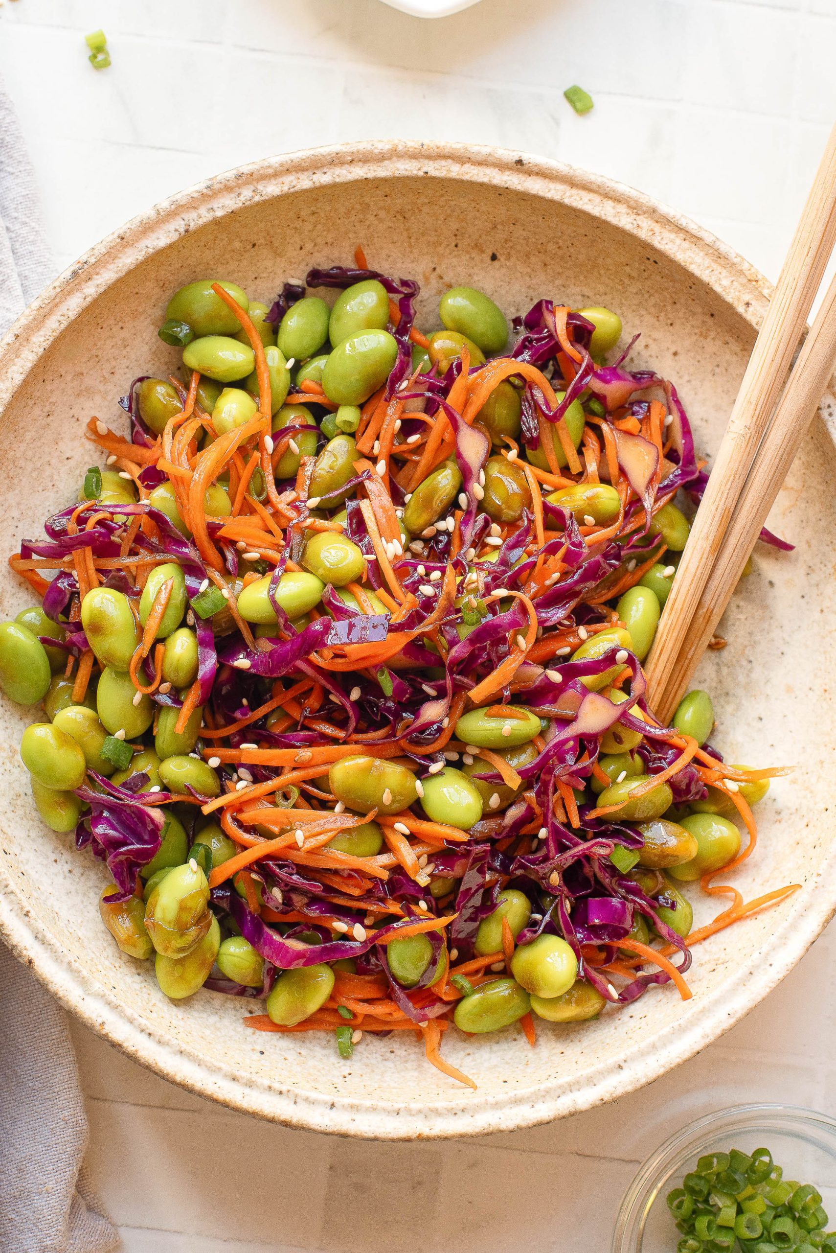 A bowl of salad with edamame, shredded carrots, red cabbage, and sesame seeds, mixed with wooden chopsticks.