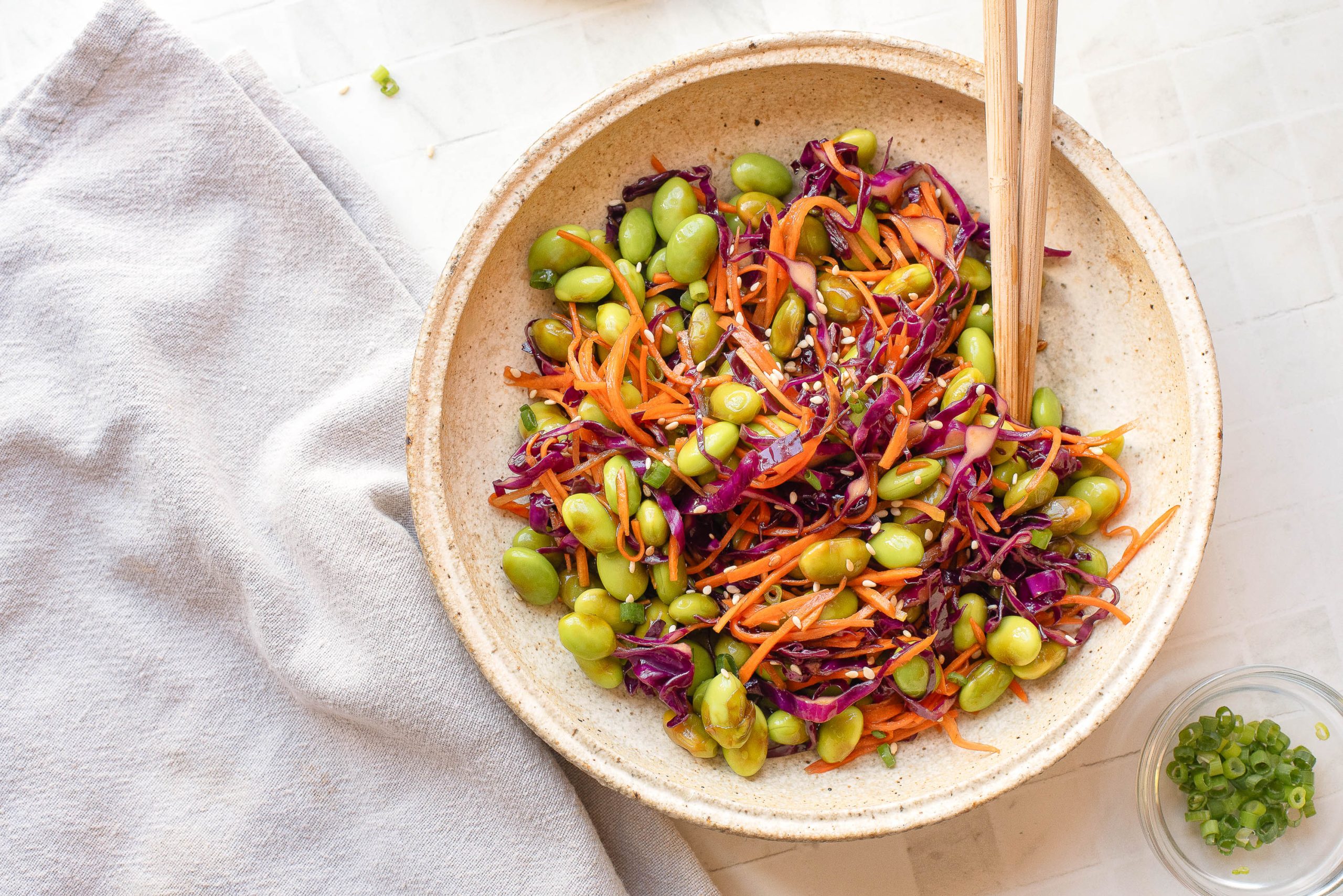 A bowl of salad with edamame, shredded carrots, and purple cabbage, garnished with sesame seeds and served with chopsticks on a white surface.