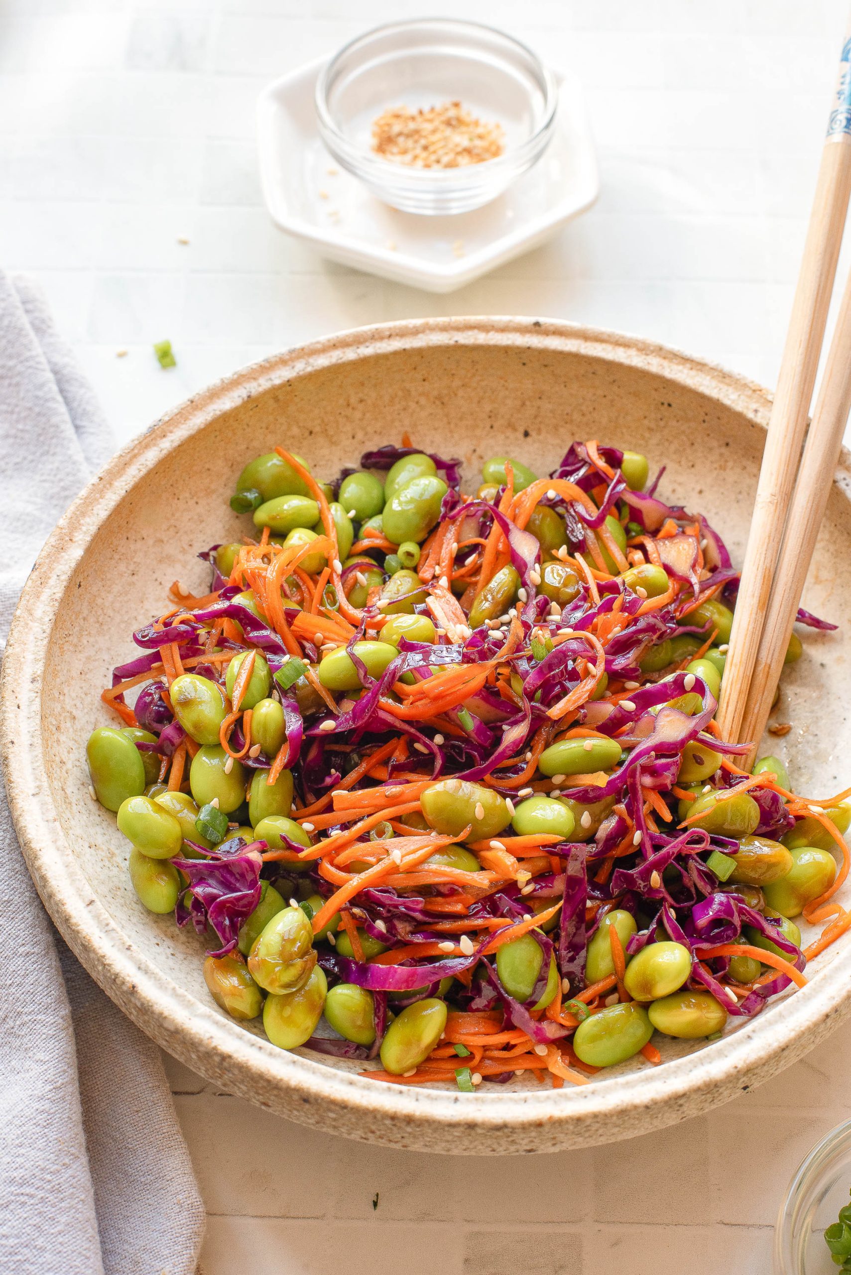 A bowl of vegetables with chopsticks placed on the side and a small dish of sesame seeds in the background.