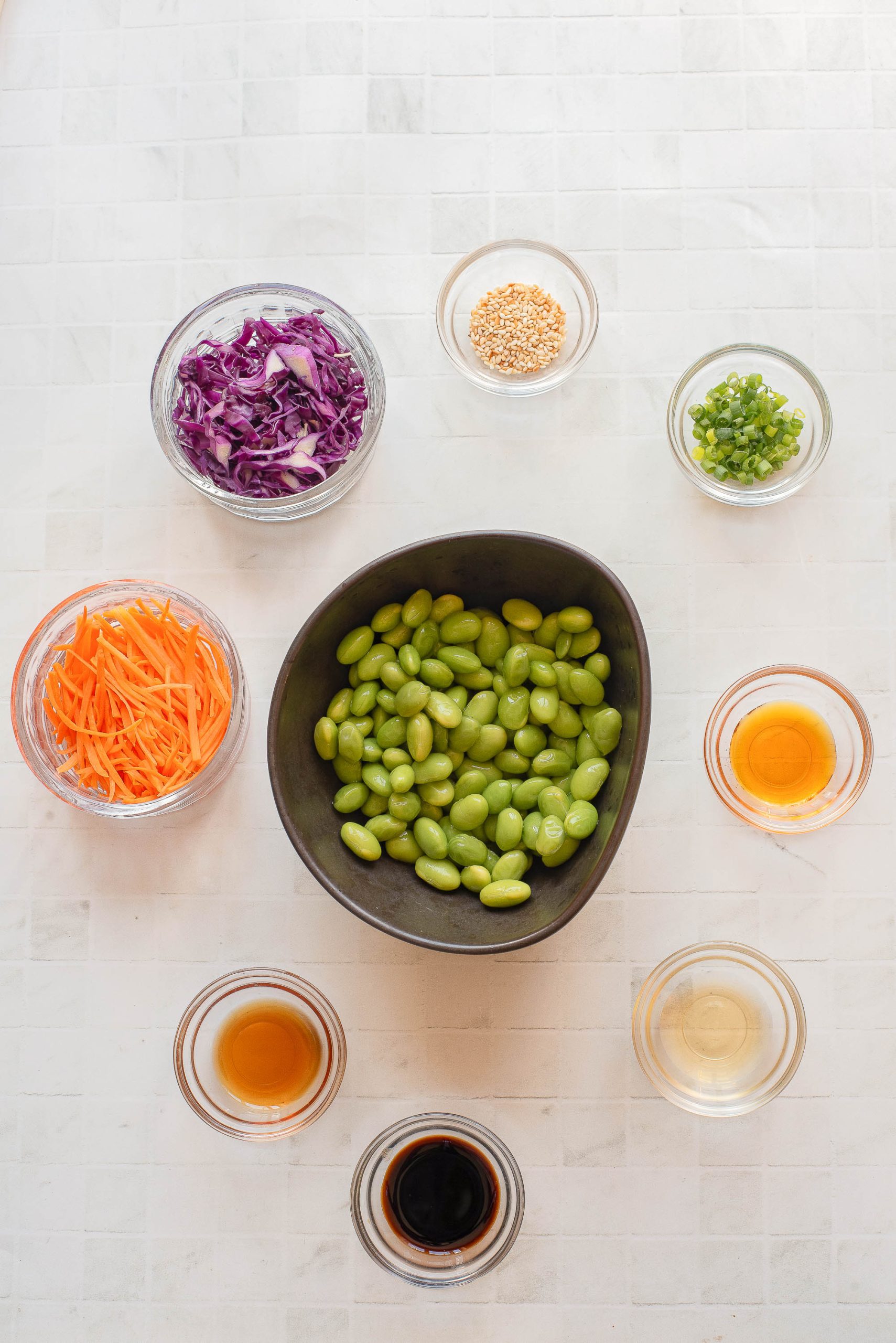 A flat lay of bowls containing edamame, shredded carrots, chopped red cabbage, green onions, sesame seeds, and various sauces on a light surface.