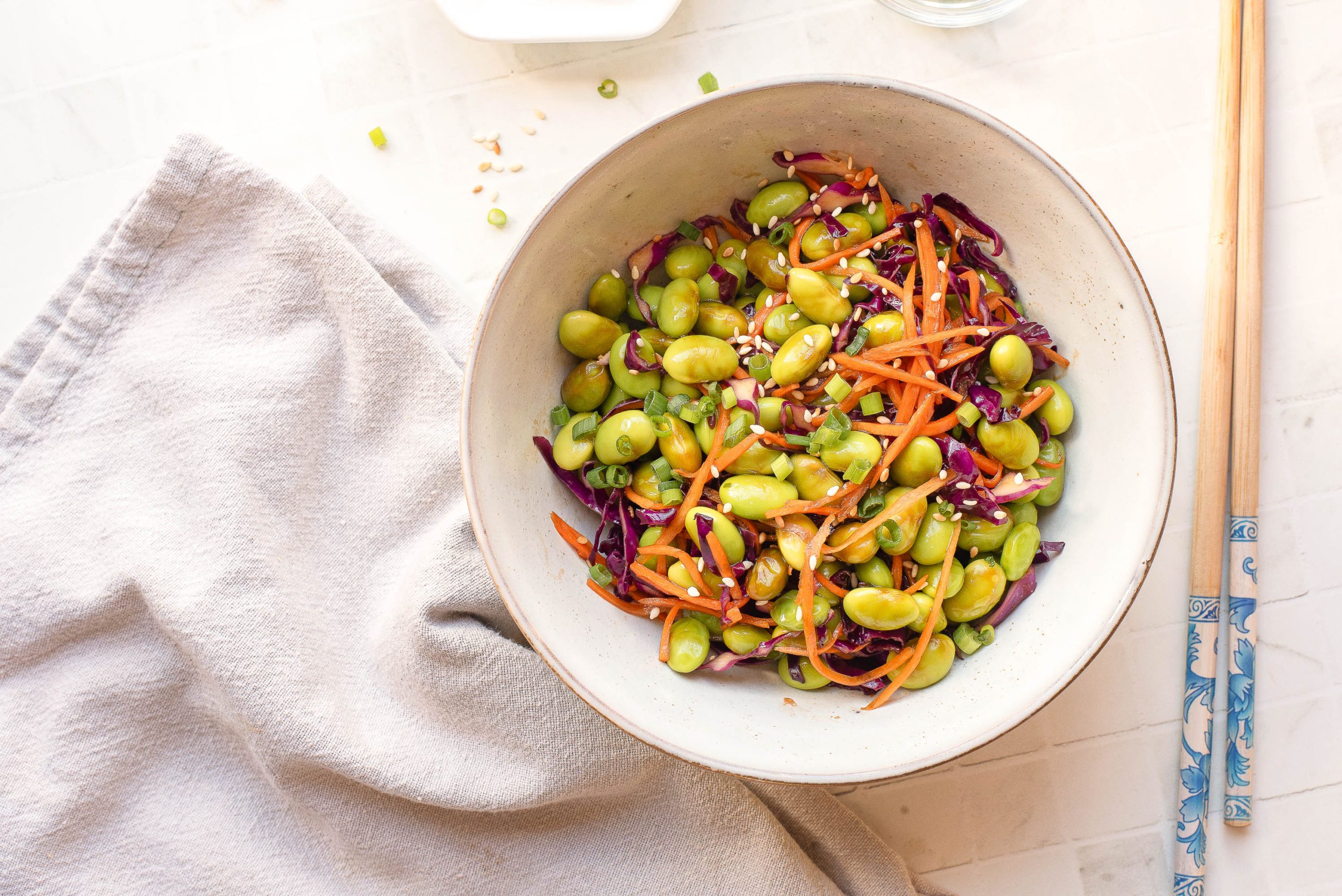 A bowl of edamame salad with shredded carrots, red cabbage, and sesame seeds sits on a light surface next to chopsticks and a folded beige cloth.