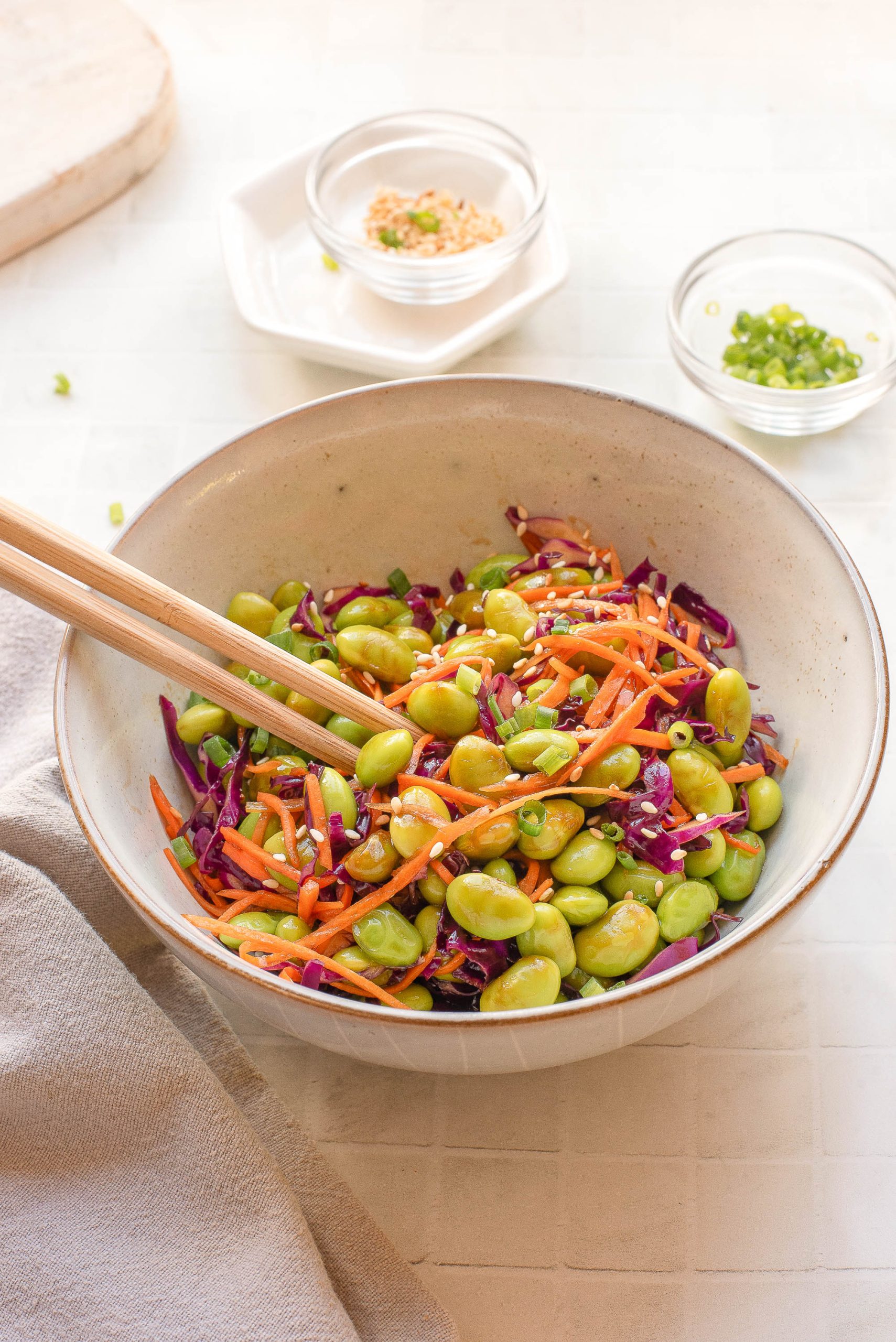A bowl of salad with edamame, shredded carrots, and purple cabbage, mixed with chopped green onions, with chopsticks resting on the bowl. Small bowls of toppings are in the background.