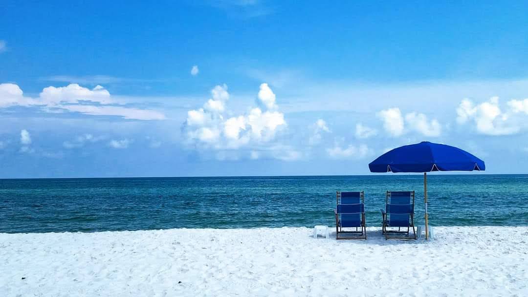 Two empty beach chairs sit under a blue umbrella on a white sandy beach, facing a calm blue ocean. The sky above is scattered with soft, drifting clouds.
