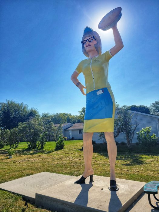 A large roadside statue of a woman in glasses, a yellow and blue uniform, and heels holds a pie aloft. The sun shines behind her head, creating a halo effect.
