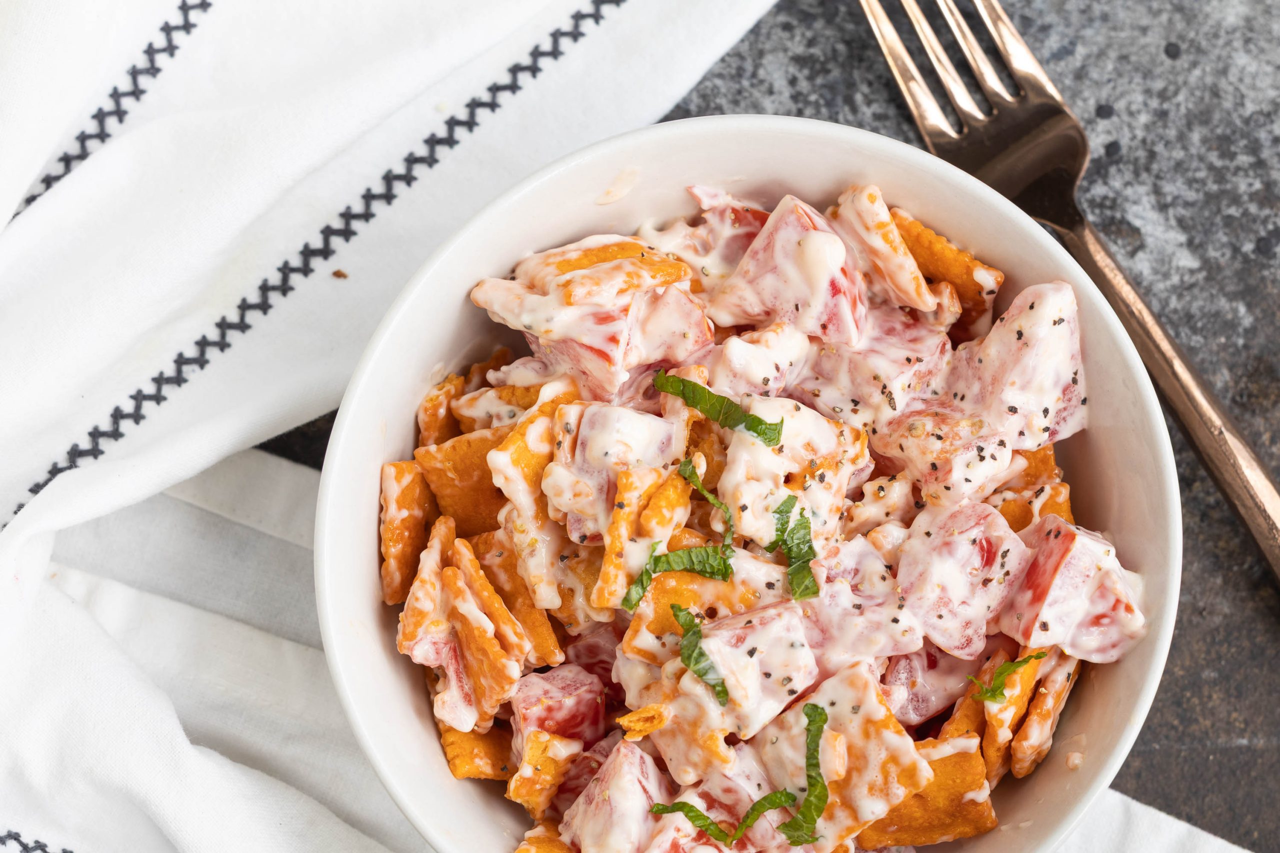 A white bowl filled with a creamy salad containing chopped tomatoes, crackers, and herbs sits on a napkin next to a fork.