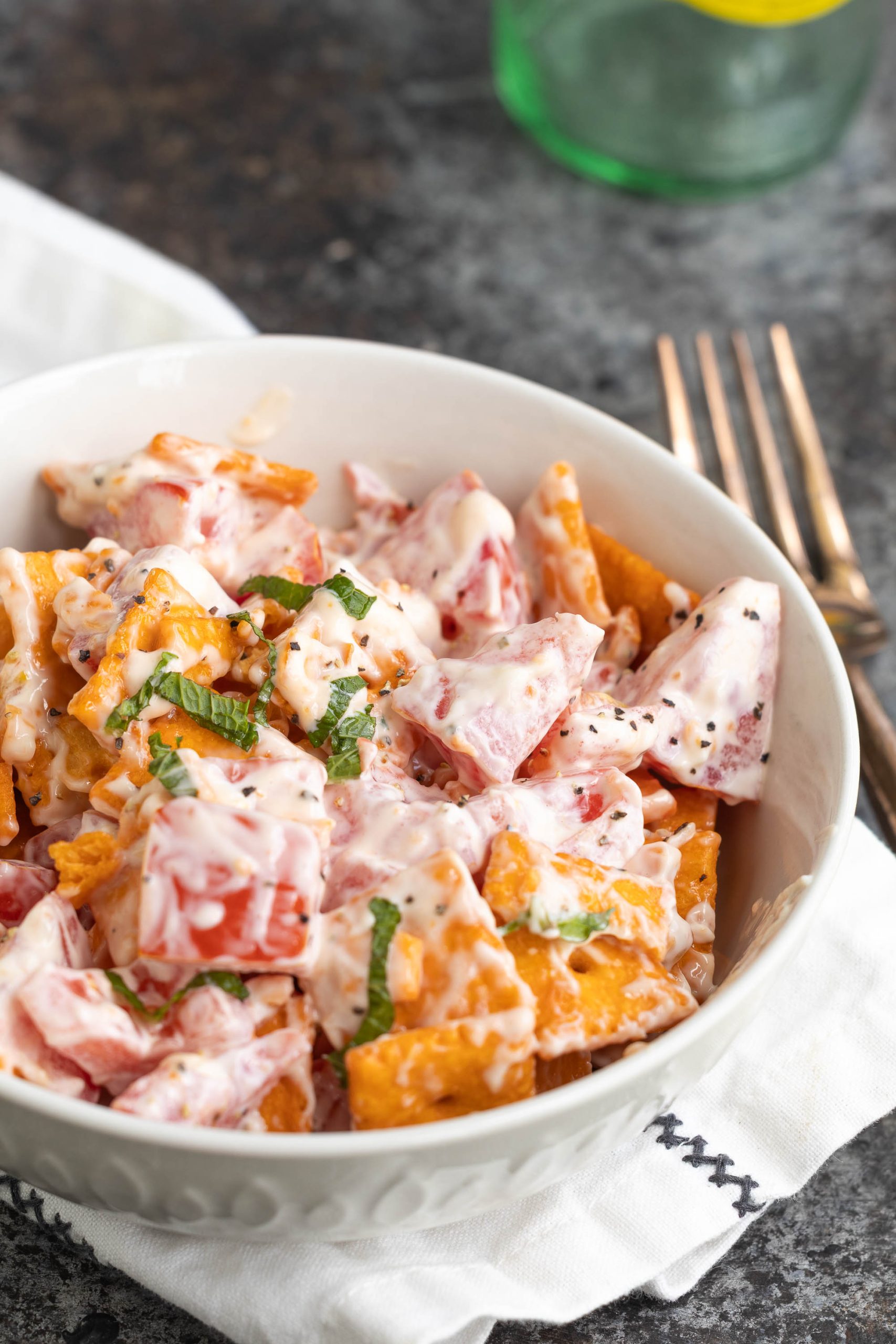 A bowl of creamy tomato and cracker salad garnished with chopped herbs, placed on a white cloth with two forks beside it.