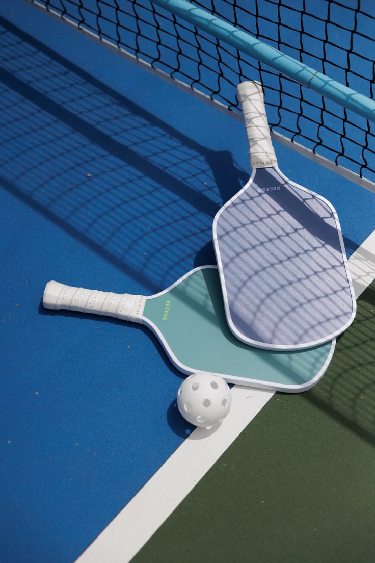 Two pickleball paddles and a perforated ball from Paula's Picks rest on a blue and green court near the net and white boundary line.