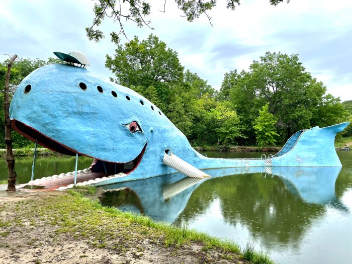A large blue whale-shaped structure with an open mouth sits partly in a pond, surrounded by trees; it has a white hat and painted smiles.