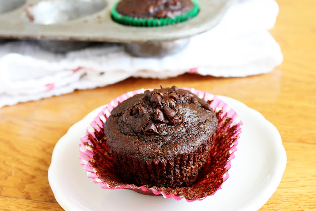 A chocolate muffin with chocolate chips sits on a white plate in a pink-striped paper liner; it's truly 50 Shades of Chocolate, with a muffin tin in the background.