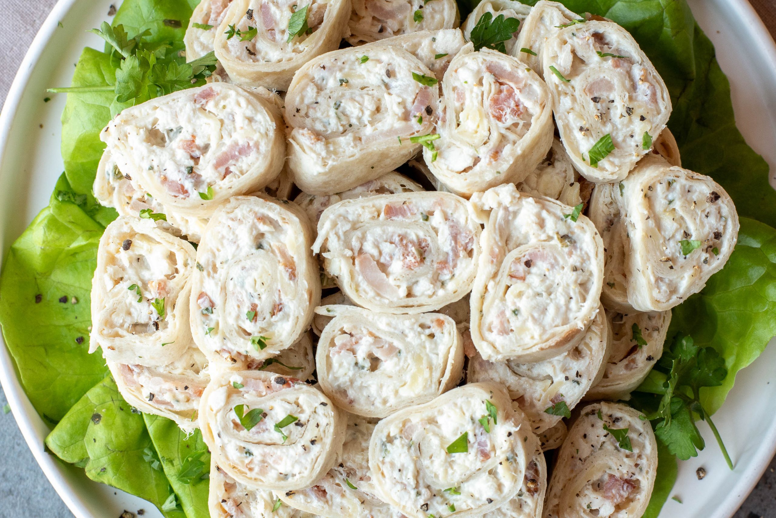 A plate of tortilla pinwheels filled with a creamy mixture, diced ham, and herbs, arranged on a bed of leafy greens.