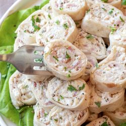 A plate of tortilla pinwheels filled with a creamy mixture, garnished with chopped parsley, served on a bed of lettuce. A fork is picking up one pinwheel.
