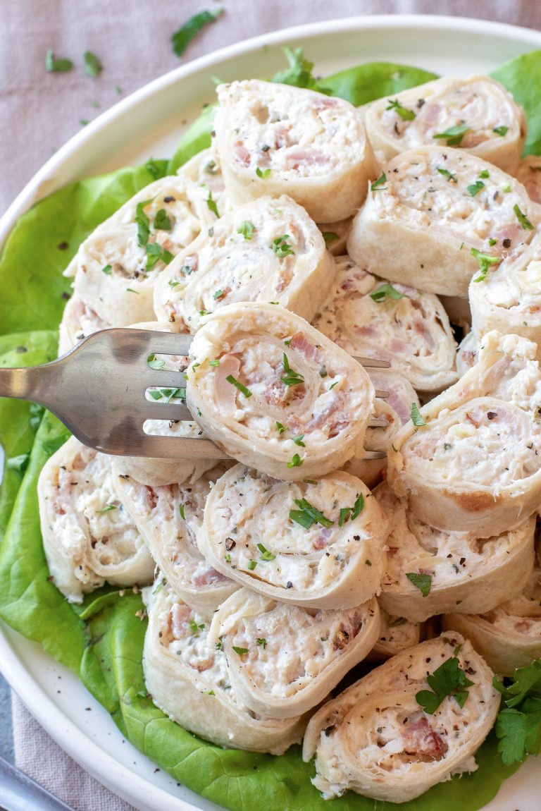 A plate of tortilla pinwheels filled with a creamy mixture, garnished with chopped parsley, served on a bed of lettuce. A fork is picking up one pinwheel.
