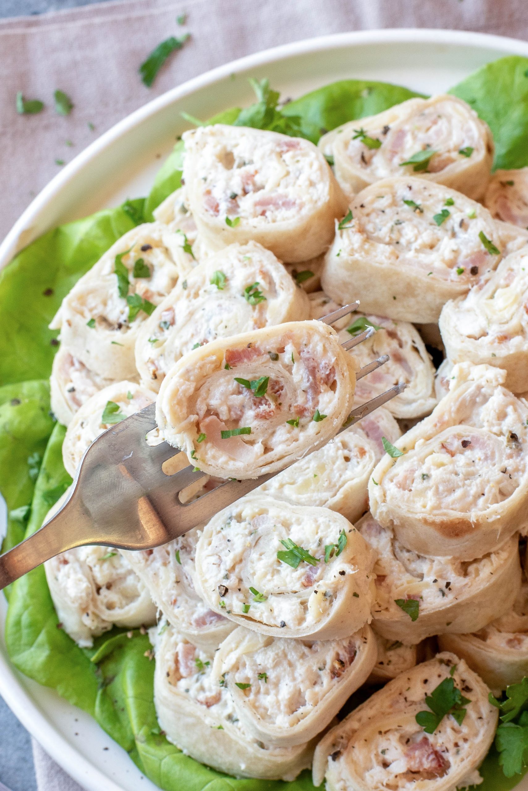 A plate of Chicken Bacon Ranch Pinwheels filled with a creamy mixture and sprinkled with herbs, served on a bed of lettuce; one roll is held up by a fork.
