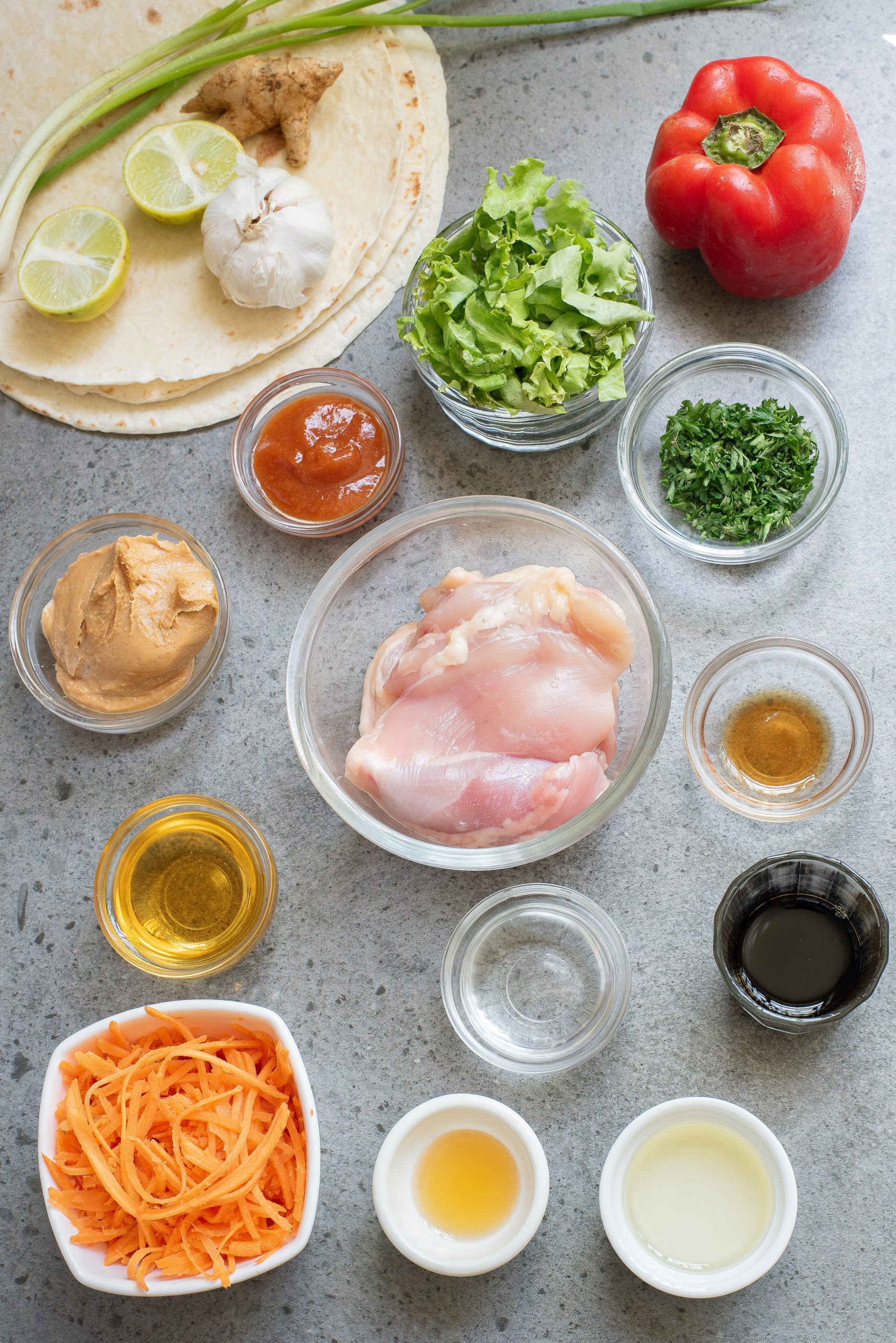 Various ingredients for a chicken wrap recipe, including raw chicken, vegetables, sauces, seasonings, and tortillas, arranged on a gray countertop.