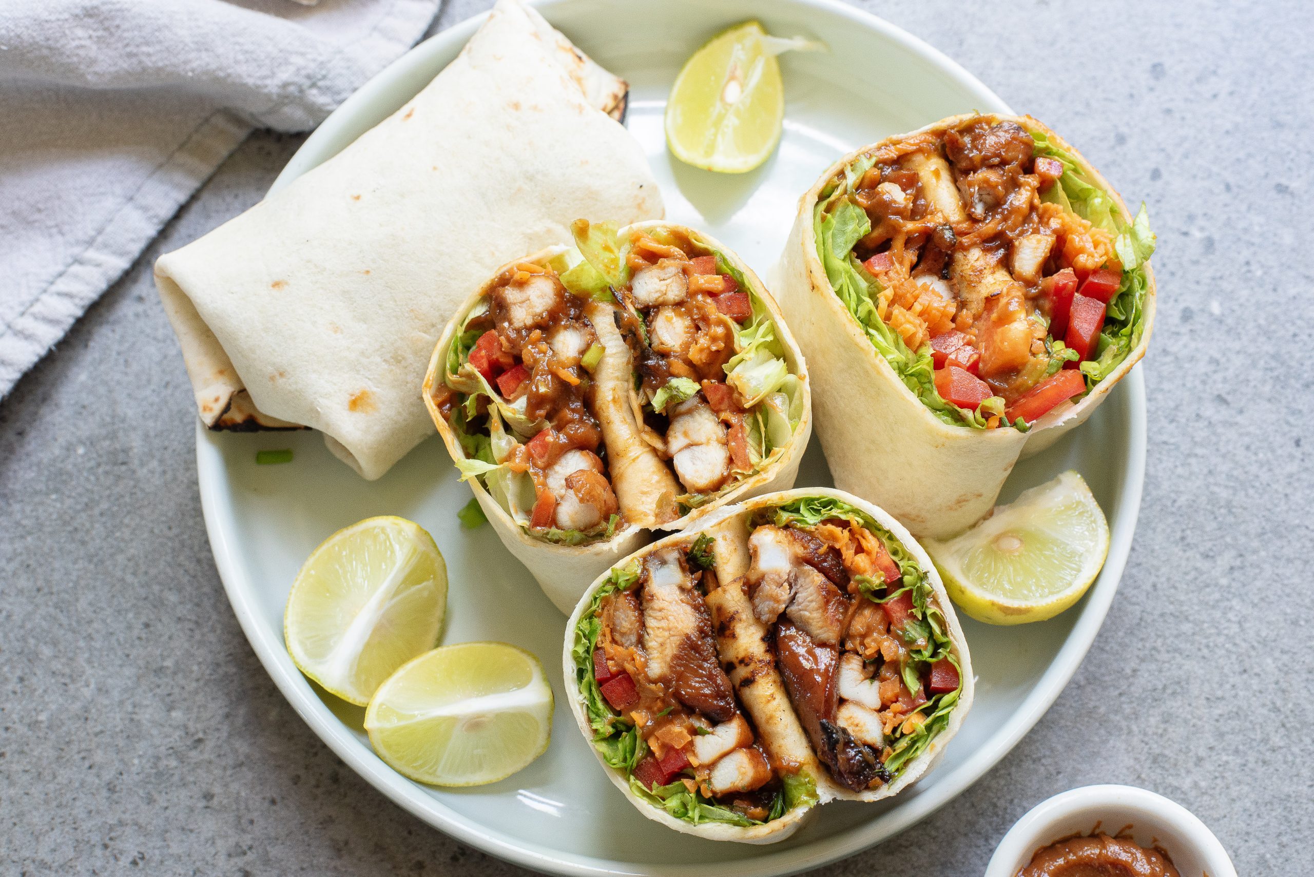 A plate with These stovetop 15-minute Asian chicken wraps are quick, easy, and full of flavor. The chicken cooks fast on the stove, and the sauce comes together with pantry basics. They're great for busy weeknights when you need something fast but better than takeout. filled with lettuce, tomato, and sauce, served with lime wedges and a whole wrap in the background.