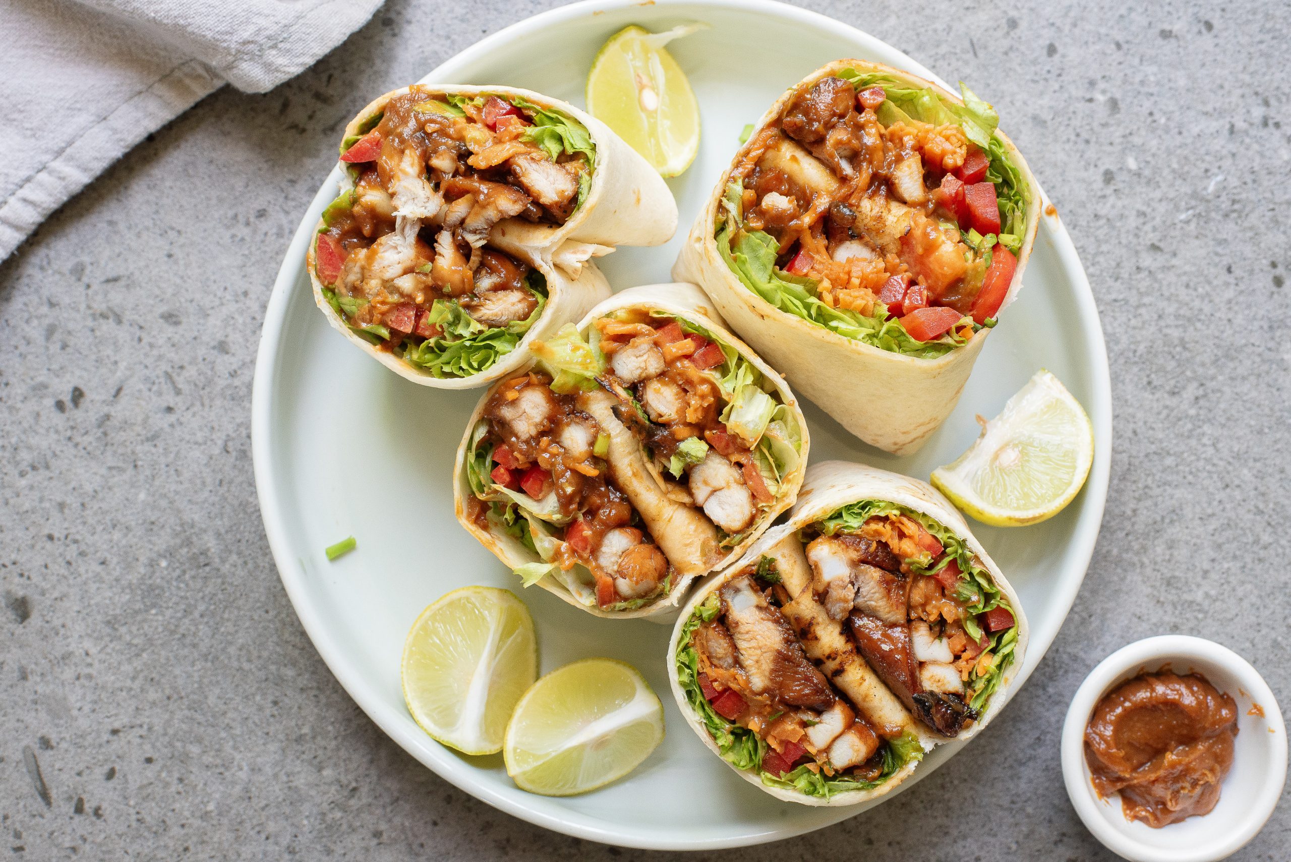 A plate with four sliced chicken wraps filled with vegetables and sauce, garnished with lime wedges, next to a small dish of dipping sauce.