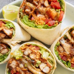 A plate with several halved chicken wraps filled with lettuce, diced tomatoes, grilled chicken, shredded cheese, and sauce, with a lemon wedge on the side.