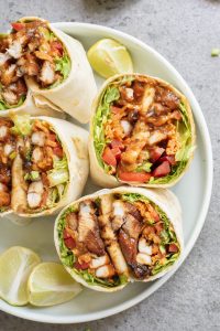 A plate with sliced chicken wraps filled with lettuce, tomatoes, and sauce, served with lime wedges on the side.
