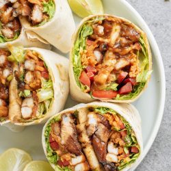 A plate with sliced chicken wraps filled with lettuce, tomatoes, and sauce, served with lime wedges on the side.
