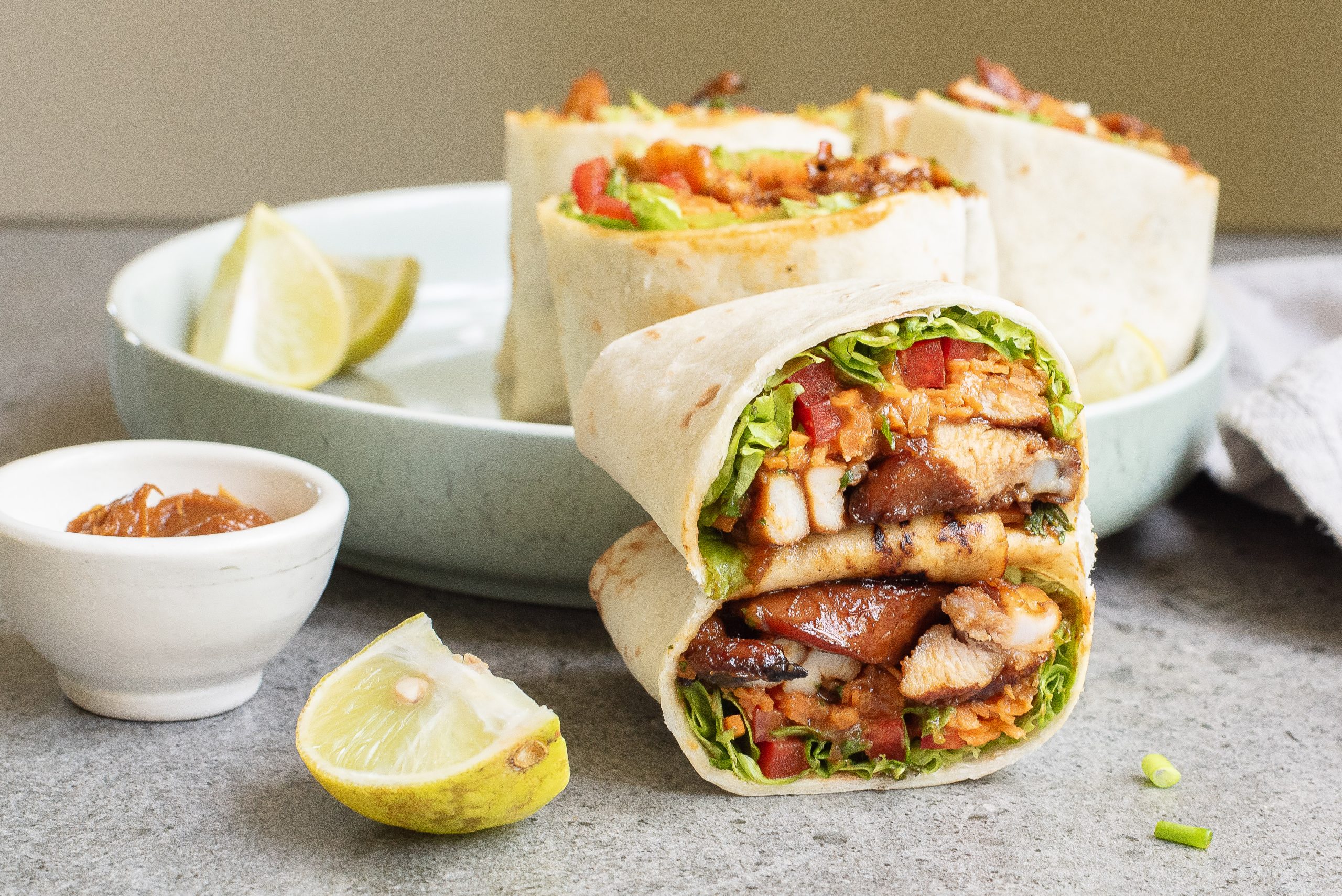 Two Stovetop 15-minute Asian Chicken Wraps are stacked on a plate, with lime wedges and a small bowl of sauce beside them.