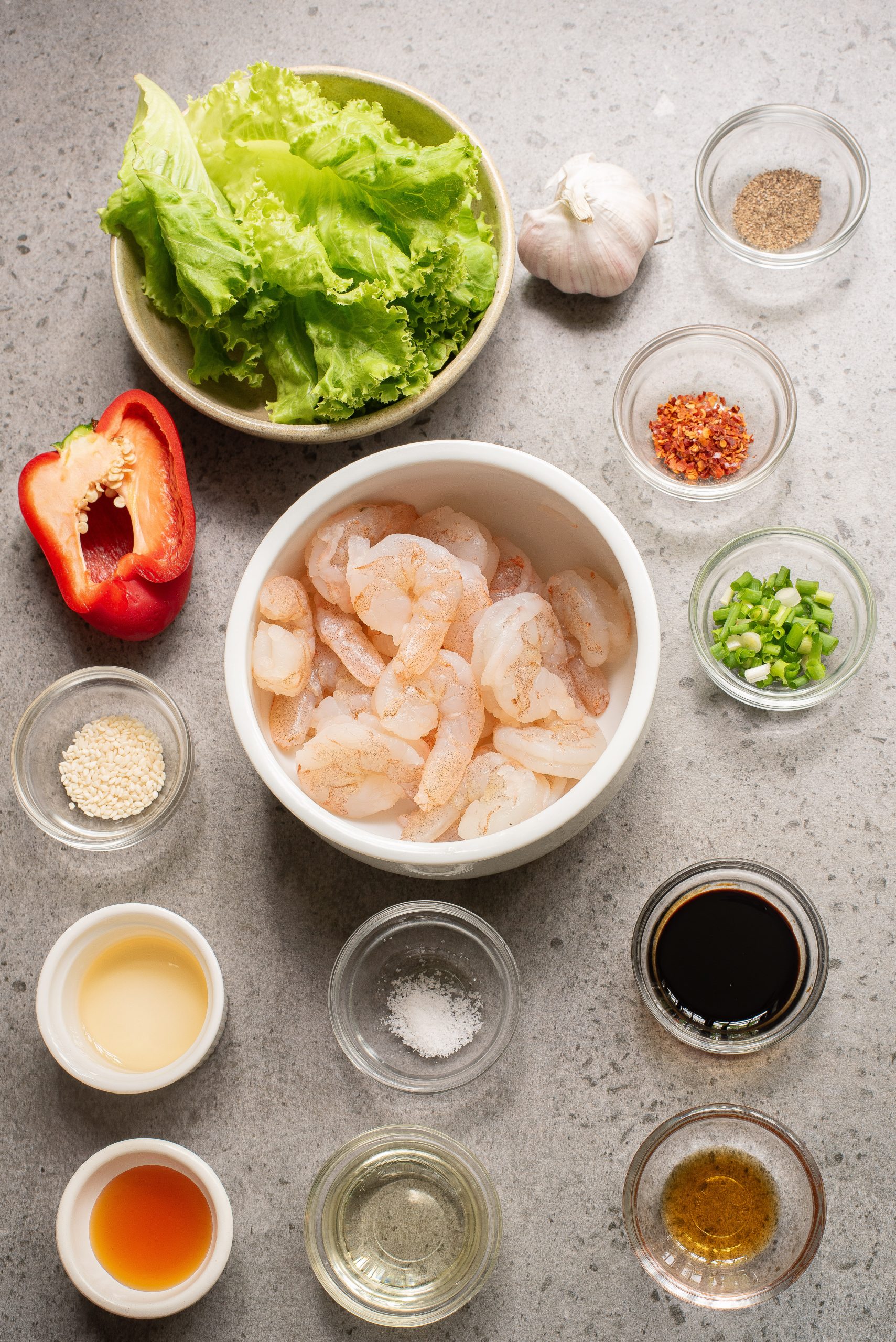 A flat lay of raw shrimp, lettuce, red bell pepper, garlic, and various spices and sauces in small bowls on a gray countertop.