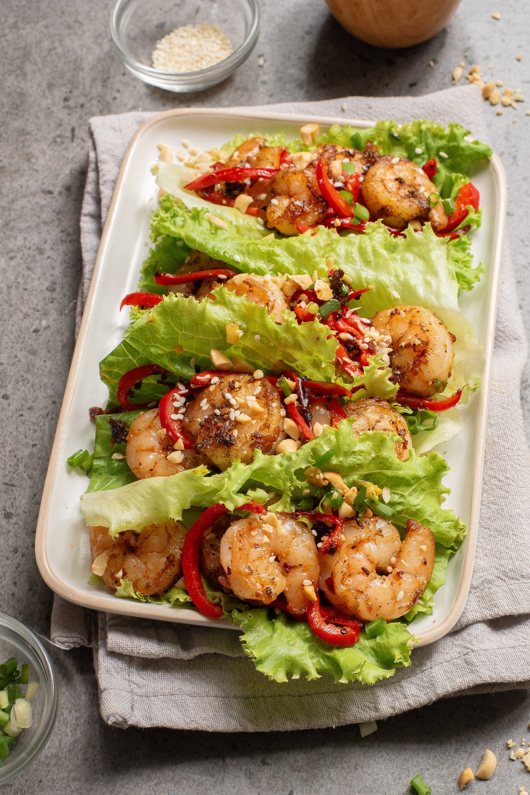 A rectangular plate with shrimp, sliced red bell peppers, and chopped peanuts, set on a gray napkin over a stone surface.