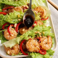 A rectangular plate with lettuce leaves topped with shrimp, red bell pepper strips, chopped peanuts, and a spoon drizzling dark sauce over the dish.