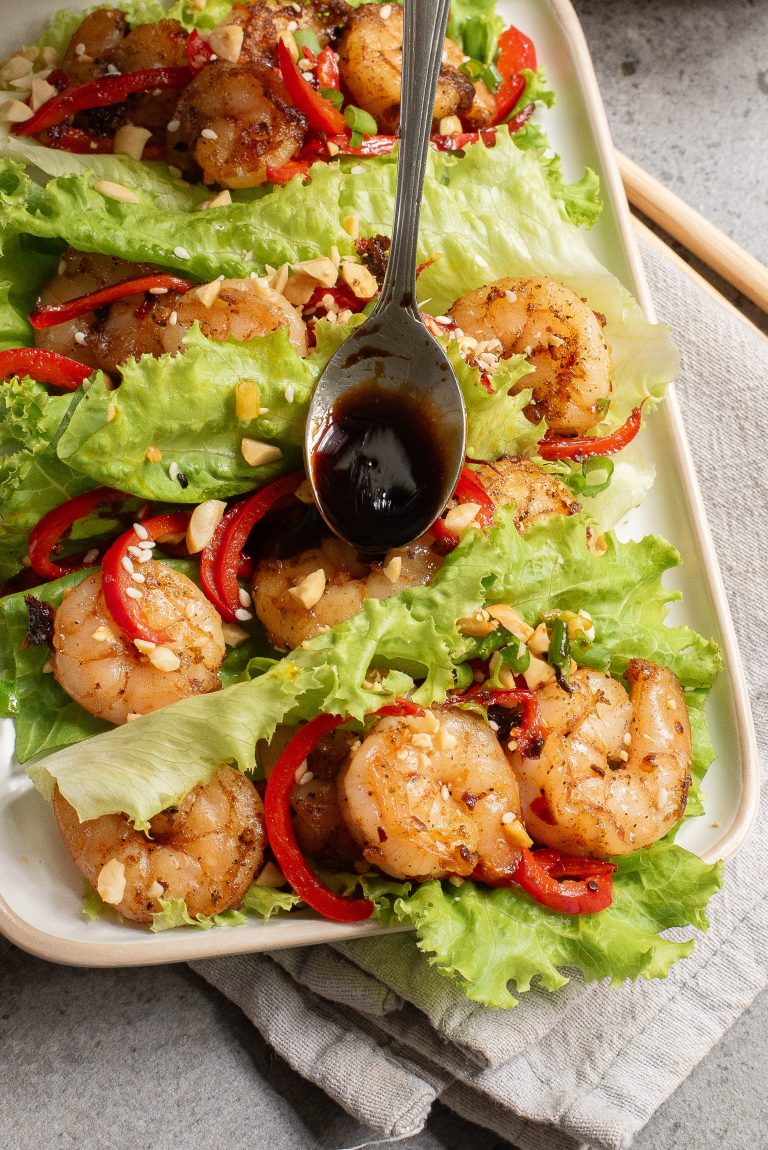 A rectangular plate with lettuce leaves topped with shrimp, red bell pepper strips, chopped peanuts, and a spoon drizzling dark sauce over the dish.