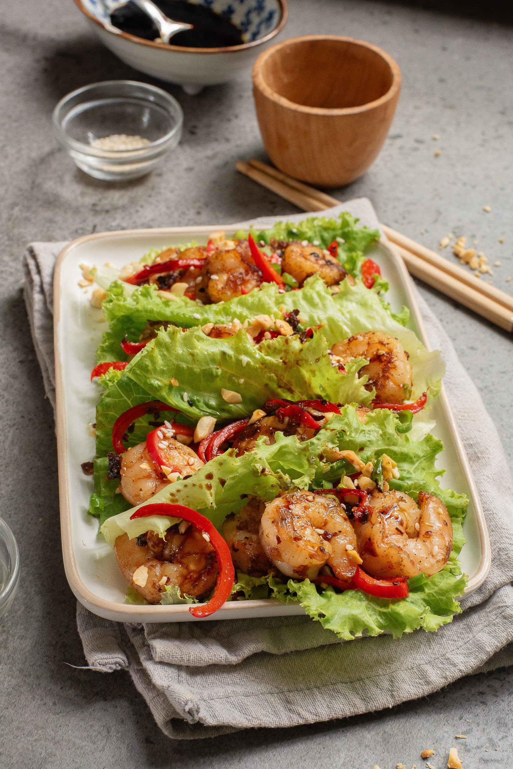 A rectangular plate with leafy green lettuce wraps filled with grilled shrimp, sliced red peppers, chopped peanuts, and herbs on a gray surface with chopsticks and bowls nearby.