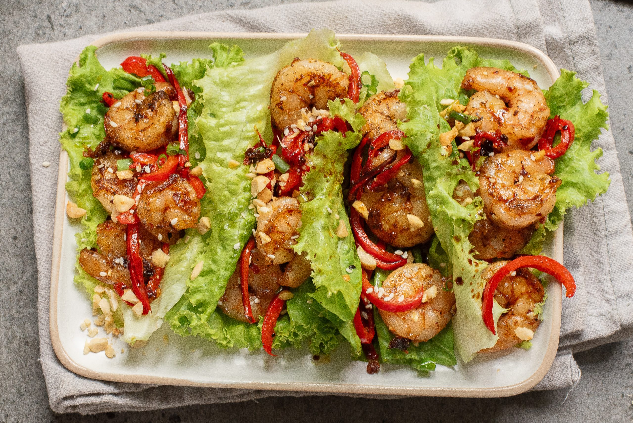 A rectangular plate with four lettuce wraps filled with cooked shrimp, sliced red peppers, chopped peanuts, and sauce, placed on a cloth napkin.
