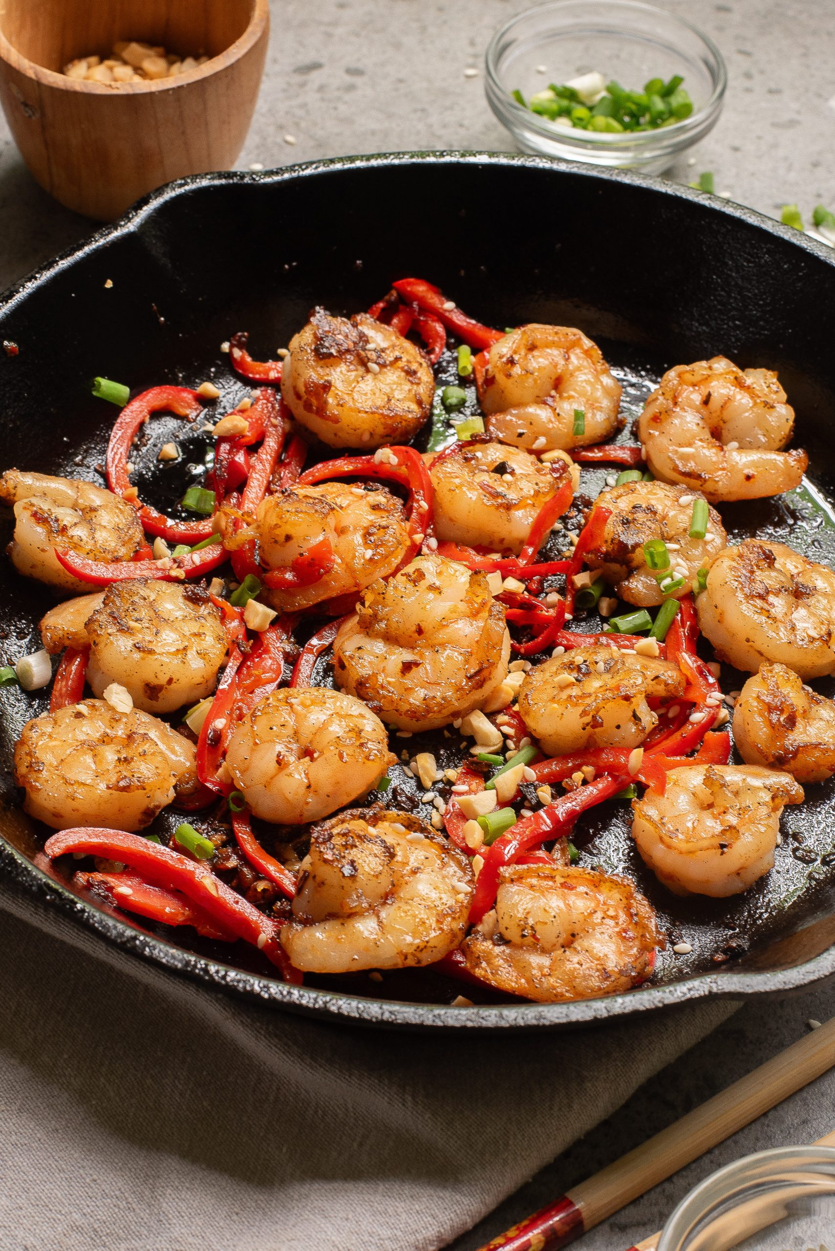 Cooked shrimp with sliced red bell peppers and chopped green onions in a black cast iron skillet, placed on a gray cloth with small bowls of condiments nearby.
