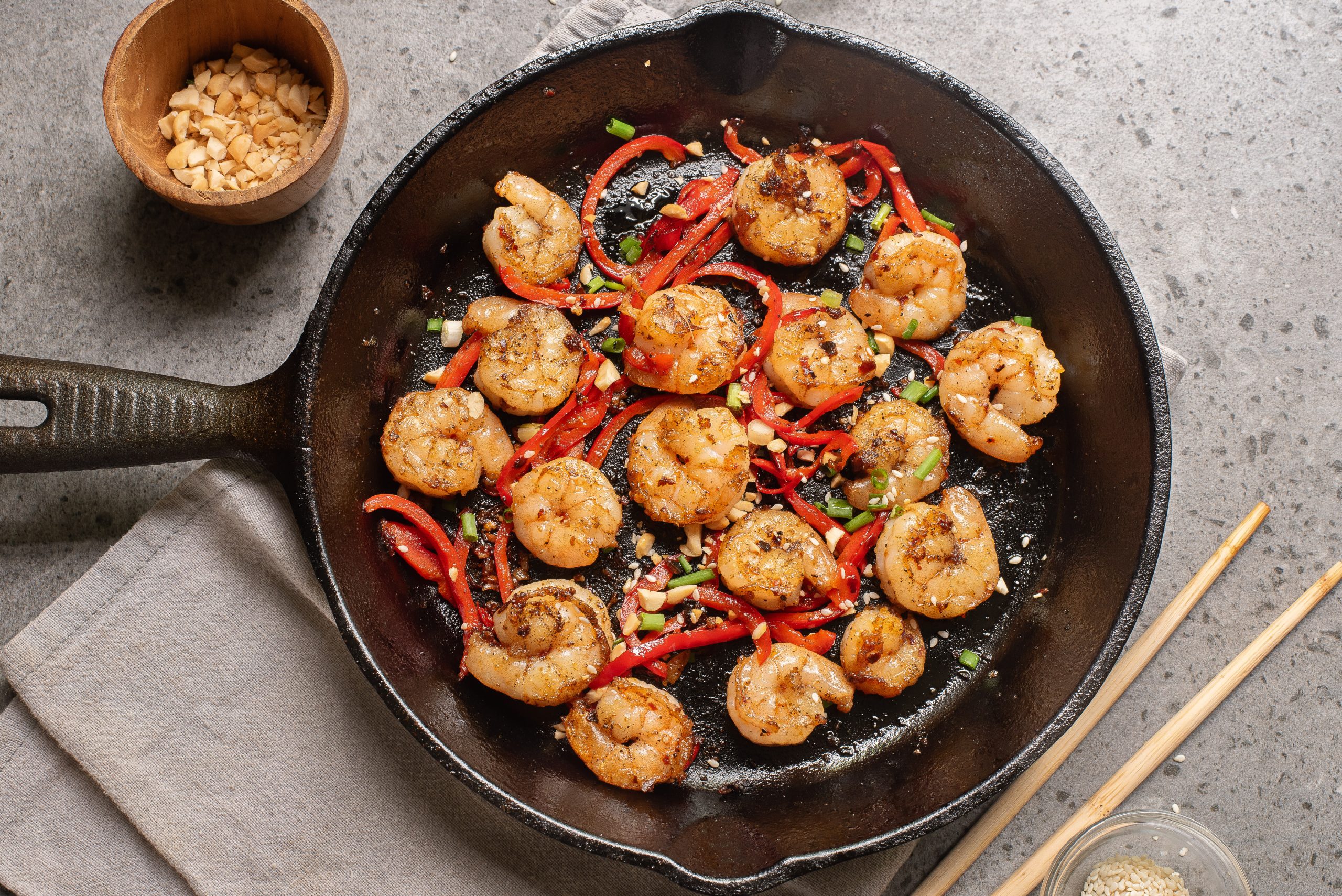 Prawns with sliced red bell pepper and green onions in a cast-iron skillet, with chopped peanuts and sesame seeds on the side.