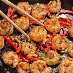 Cooked shrimp with sliced red peppers, chopped peanuts, and green onions in a cast iron skillet, with chopsticks picking up a shrimp.