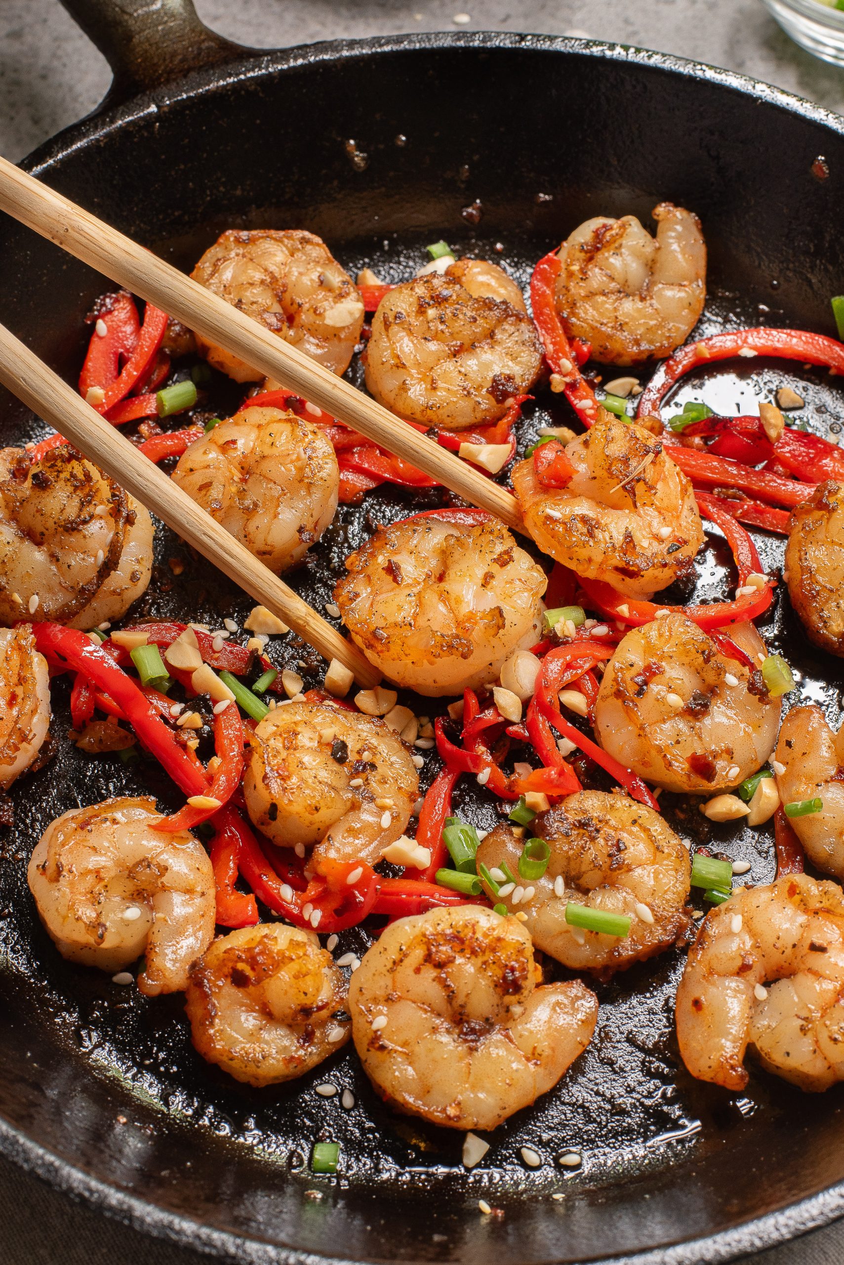 Cooked shrimp with sliced red peppers, chopped peanuts, and green onions in a cast iron skillet, with chopsticks picking up a shrimp.