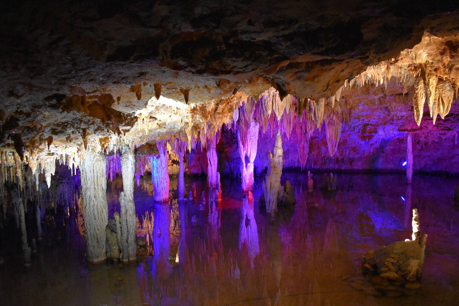 Stalactites and stalagmites are illuminated by colorful lights and reflected in the still water of an underground cave.