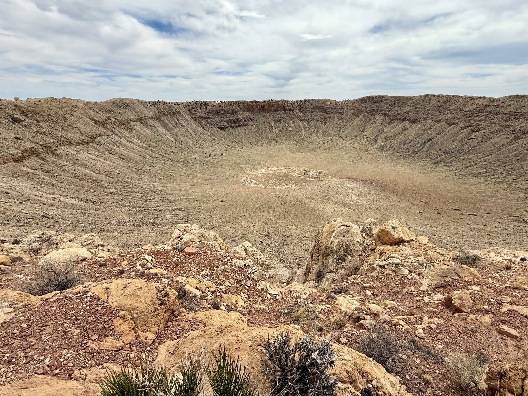 Natural Attractions of Route 66. Wide view of a large, rocky crater with a flat bottom and steep walls under a partly cloudy sky. Sparse vegetation appears in the foreground.