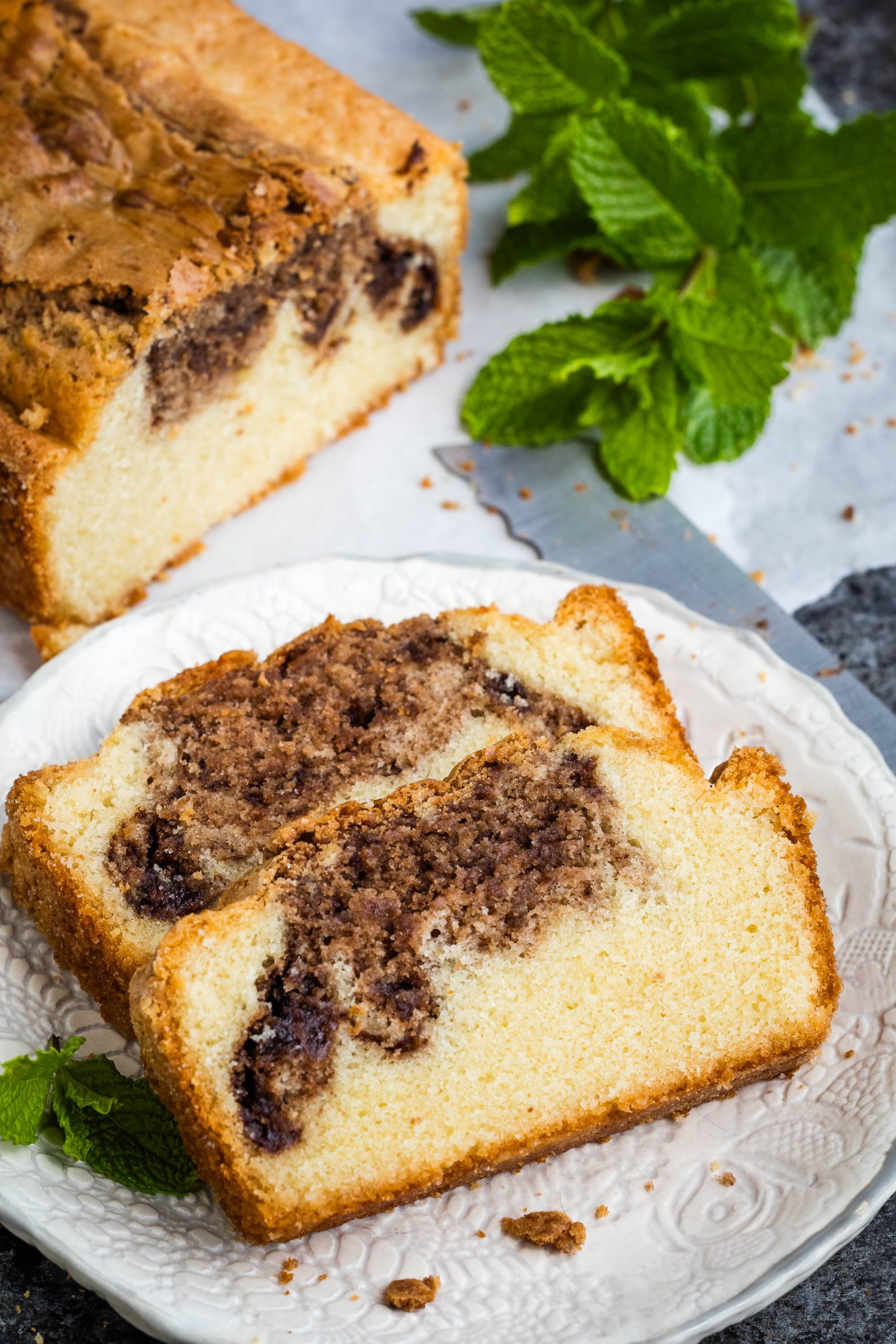 Two slices of dessert with visible chocolate swirls rest on a white plate, with the remaining loaf and mint leaves in the background.