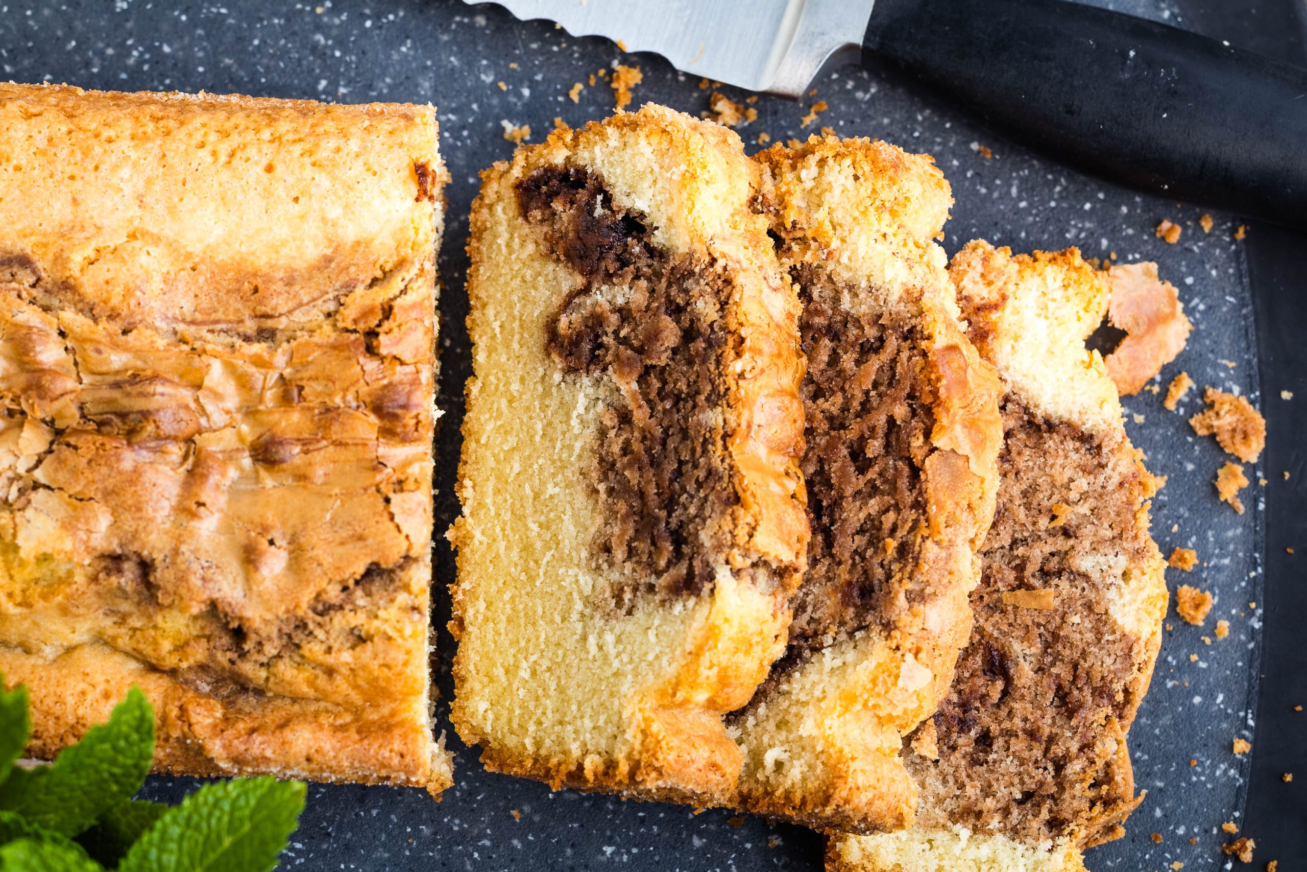 A close-up of a Nutella Marble Pound Cake, partially sliced, on a dark cutting board with a serrated knife beside it.