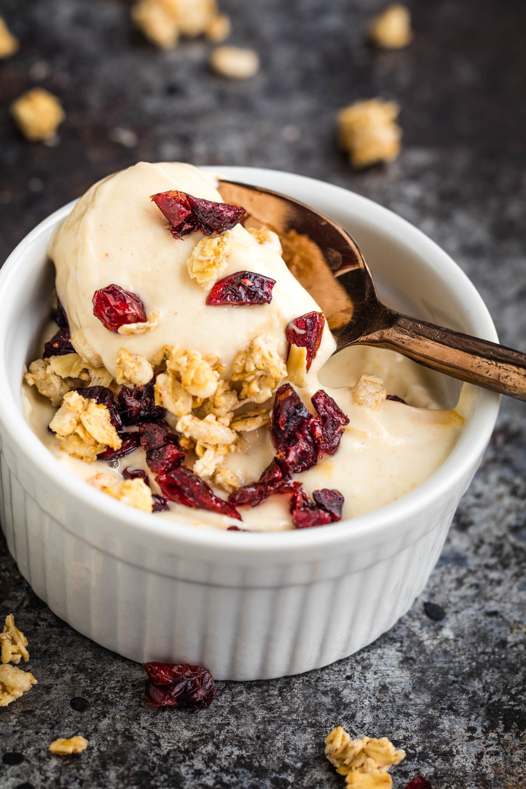 A white ramekin filled with creamy Peanut Butter Protein Greek Yogurt, topped with granola and dried cranberries, with a spoon scooping some out.