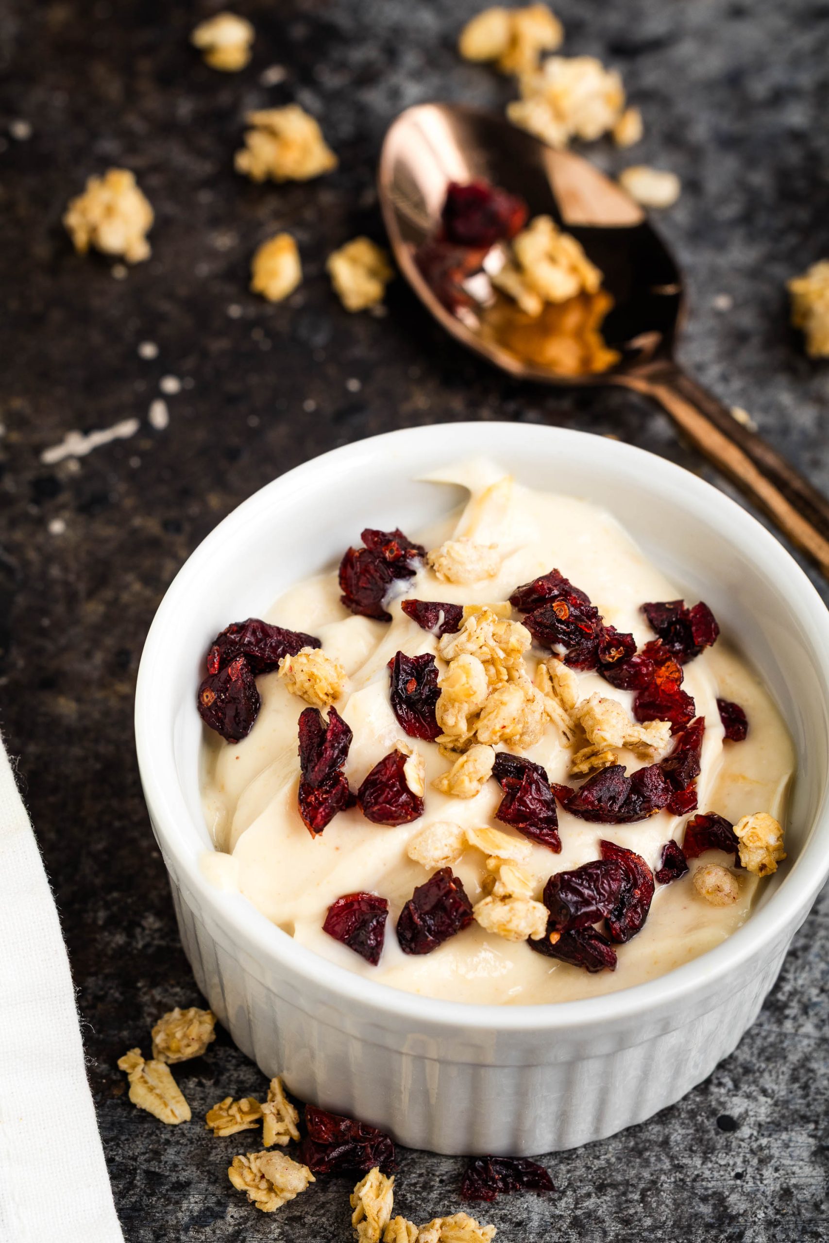 A white ramekin filled with Peanut Butter Protein Greek Yogurt topped with granola and dried cranberries sits on a dark surface; a spoon with more granola rests nearby.