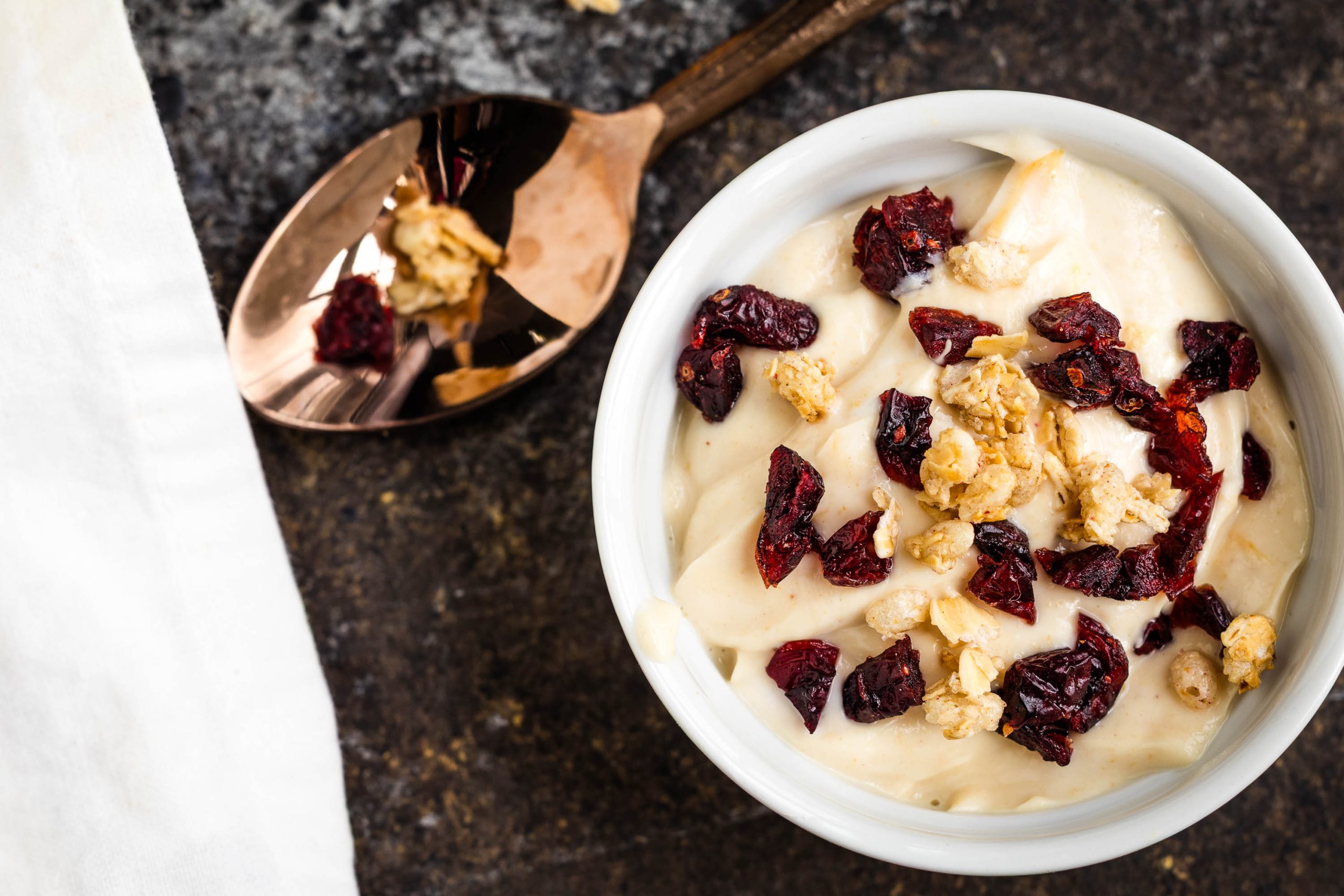 A bowl of creamy yogurt topped with granola and dried cranberries sits next to a spoon on a dark surface.