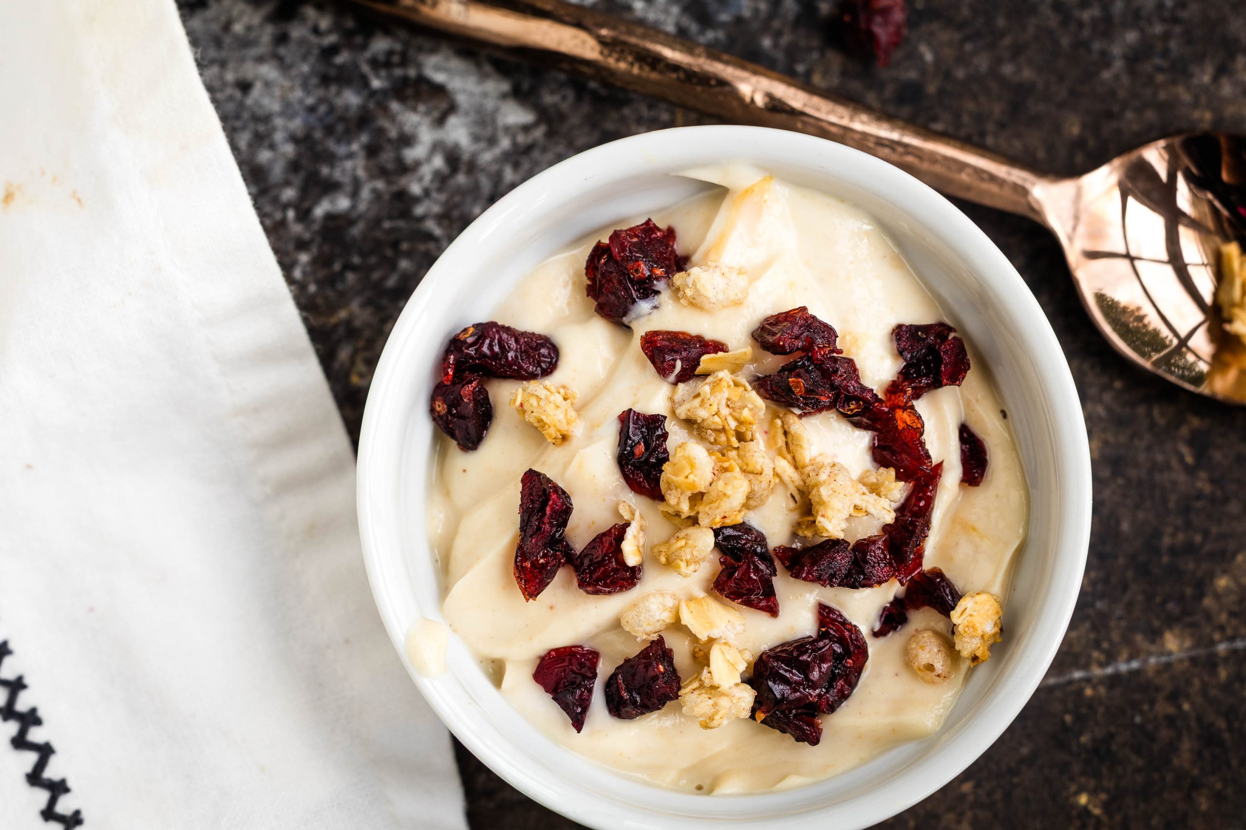 A bowl of creamy yogurt topped with granola and dried cranberries sits next to a bronze spoon on a dark surface.
