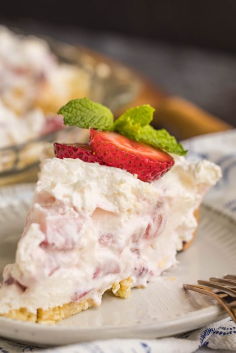 A slice of strawberry cream pie topped with whipped cream, fresh strawberry slices, and a mint sprig sits on a plate beside a fork.