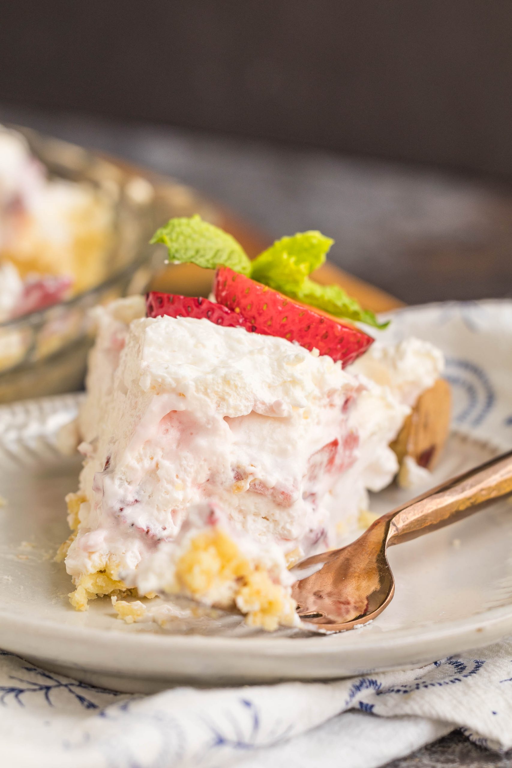 A slice of creamy dessert with layers of whipped topping, sliced strawberries, and a garnish of mint, served on a plate with a fork.