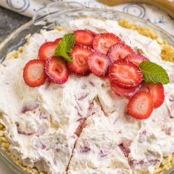 A pie with a crumbly crust, topped with whipped cream, sliced strawberries, and mint leaves, sits partially sliced on a glass pie plate.