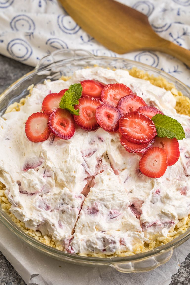 A pie with a crumbly crust, topped with whipped cream, sliced strawberries, and mint leaves, sits partially sliced on a glass pie plate.