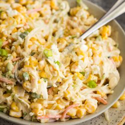 A bowl of creamy corn salad with shredded carrots, green onions, and a fork, on a speckled plate.